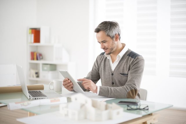 A man working on his Ipad with his laptop, coffe, phone and glasses on the table